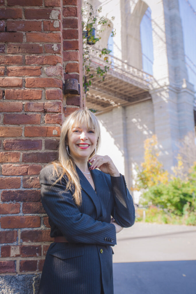 woman in a suit, smiling, leaning against brick wall.